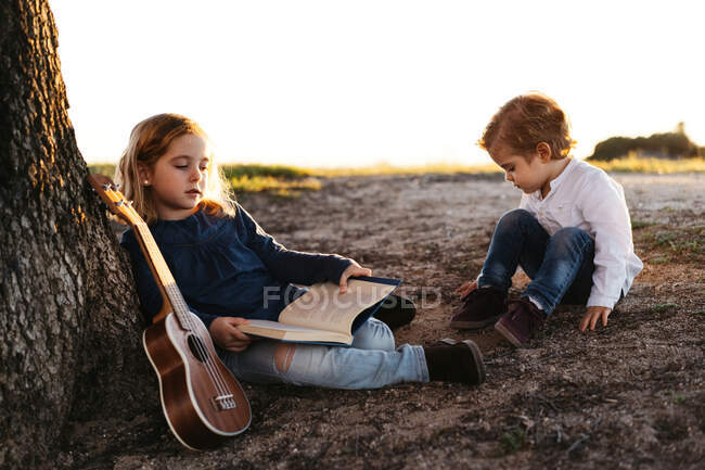 Side view of adorable little girl reading interesting story to younger brother while sitting together under tree with ukulele guitar in summer day in countryside — Stock Photo