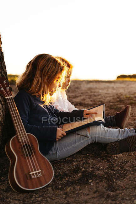 Літній день у сільській місцевості перед молодшим братом відкривається цікавий краєвид на маленьку дівчинку, яка сидить разом під деревом з гітарою укулеле. — стокове фото