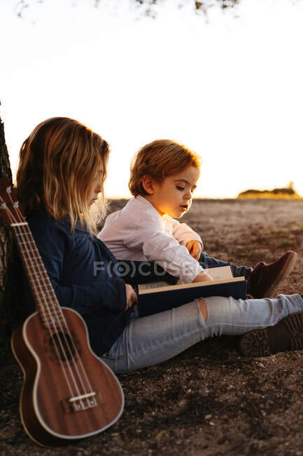 Seitenansicht des entzückenden kleinen Mädchens lesen interessante Geschichte zu jüngeren Bruder, während sie zusammen unter Baum mit Ukulele-Gitarre im Sommertag in der Natur sitzen — Stockfoto