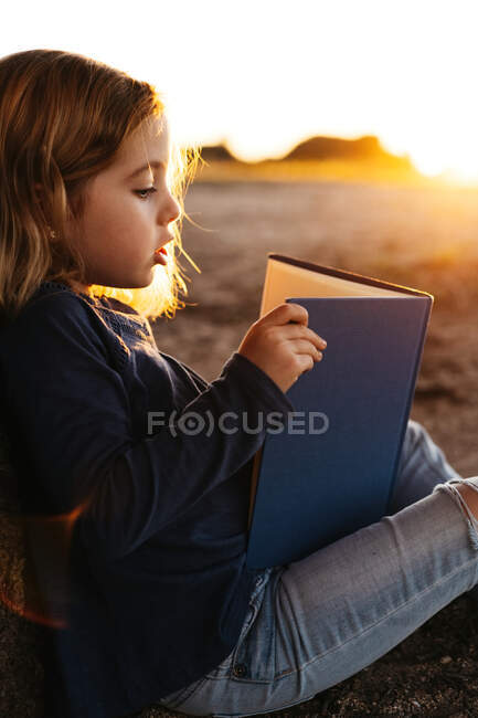 Vista lateral da menina calma no desgaste casual leitura de conto de fadas livro enquanto sentado sob a árvore em campo na noite de verão ensolarado — Fotografia de Stock