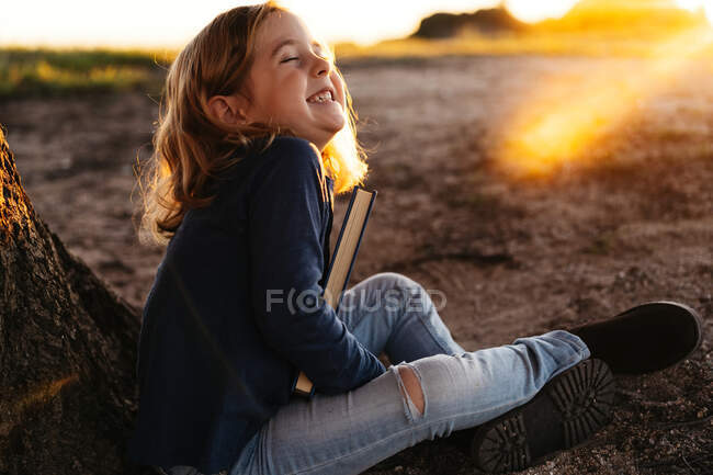 Side view of calm little girl with closed eyes smiling holding a fairy tale book while sitting under tree in field in sunny summer evening — Stock Photo