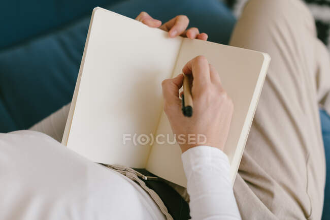Dall'alto donna senza volto in camicia bianca seduta sul divano e scritta nel taccuino che lavora sul progetto aziendale — Foto stock