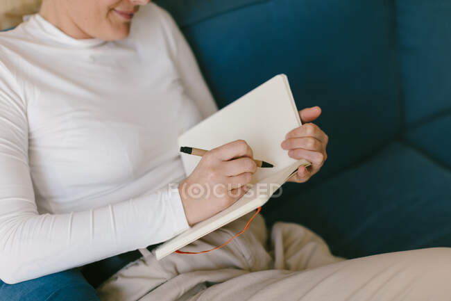Сверху безликая женщина в белой рубашке сидит на диване и пишет в блокноте, работая над бизнес-проектом — стоковое фото