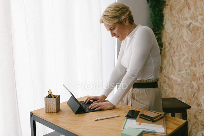 Focalisé blond adulte femme debout à la table en bois avec papeterie tapant sur clavier portable de la tablette contre la fenêtre de la lumière — Photo de stock