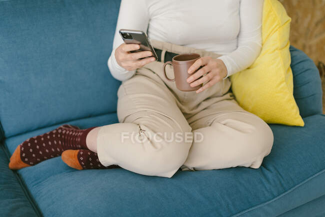 Mujer sin rostro copped sin zapatos escalofriante en acogedor sofá en la oficina disfrutando de la taza de café y el surf teléfono móvil — Stock Photo