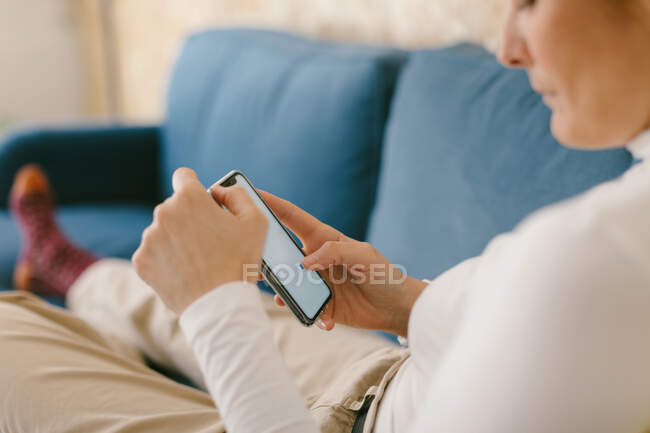 Безликая женщина без обуви отдыхает на уютном диване в офисе наслаждаясь чашкой кофе и серфингом мобильного телефона — стоковое фото