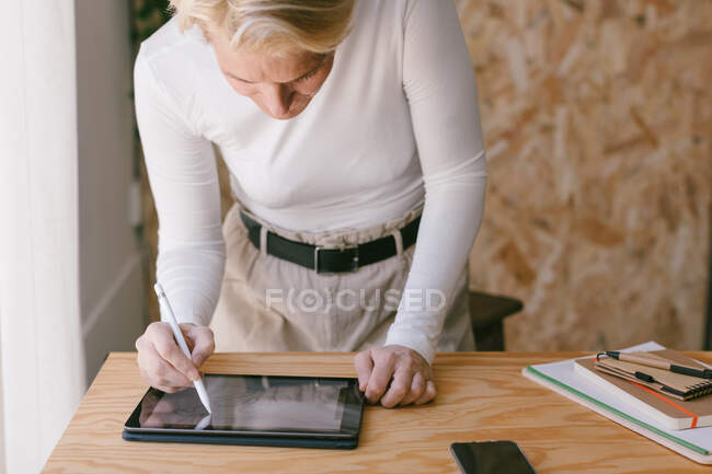 Stylish blond businesswoman bending over table and working on tablet with stylus in light wooden office — Stock Photo