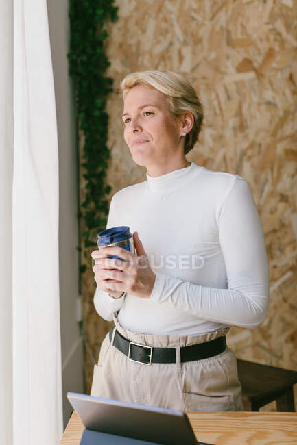 Smiling happy adult woman at working desk enjoying morning coffee while smiling away in office window — Stock Photo