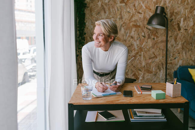 Konzentrierte erwachsene Geschäftsfrau schaut weg, während sie sich auf Klemmbrettern Notizen macht, die sich auf einem Holztisch im hellen Büro biegen. — Stockfoto