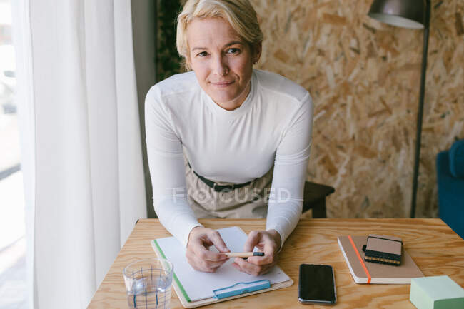 Konzentrierte erwachsene Geschäftsfrau blickt in die Kamera, während sie sich auf dem Klemmbrett Notizen macht und sich auf dem Holztisch im hellen Büro beugt — Stockfoto