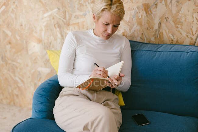 Blonde Frau mit kurzen Haaren in weißem Hemd sitzt auf Sofa und schreibt in Notizbuch an Geschäftsprojekt — Stockfoto