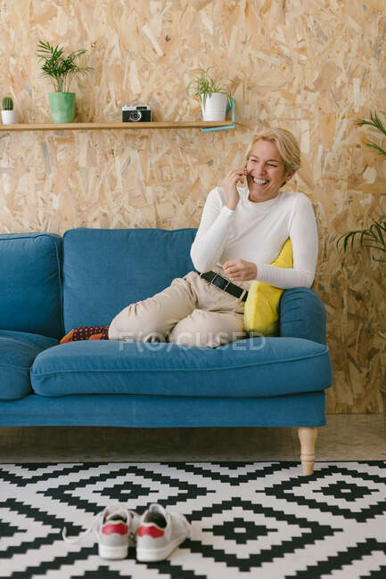 Vista laterale della donna allegra casuale che messaggia sullo smartphone mentre si interrompe in ufficio seduta sul divano — Foto stock
