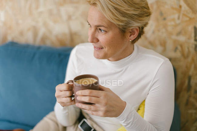 Calma mujer de negocios adulta con el pelo corto y rubio sentado en el sofá acogedor en la oficina teniendo taza de café y sonriendo tranquilamente mirando hacia otro lado - foto de stock