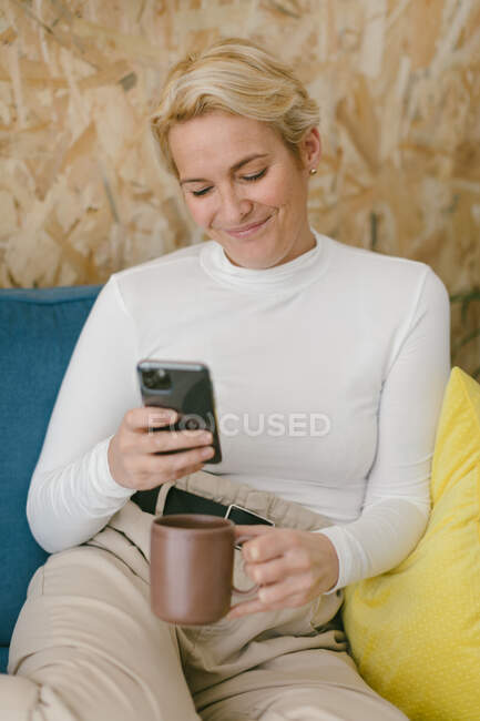 Empresária loira com cabelo curto refrigerando no sofá aconchegante no escritório desfrutando de xícara de café e surfando celular — Fotografia de Stock
