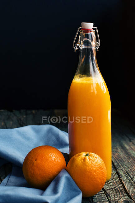 Botella de zumo de naranja cítrico fresco colocada cerca de naranjas frescas sobre una mesa rústica oscura de madera al fondo oscuro - foto de stock