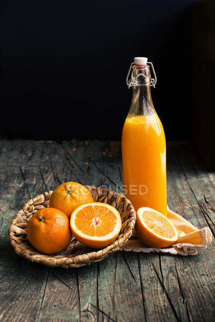 Пляшка свіжого лимонного апельсинового соку, розміщеного біля половини свіжих апельсинів на дерев'яному темному сільському столі на темному фоні — стокове фото