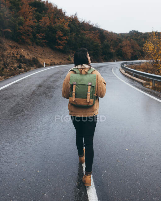 Vista posteriore del giovane escursionista femminile attivo in abito casual con zaino a piedi sulla strada asfaltata bagnata vuota che conduce attraverso la foresta colorata nella giornata autunnale — Foto stock