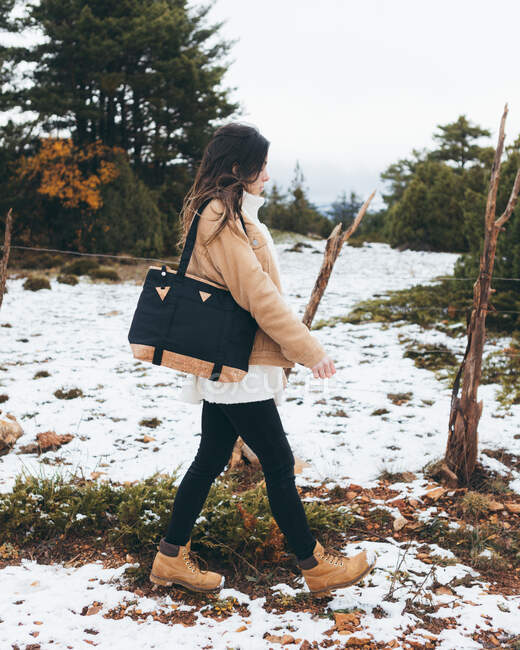 Side view of young active female hiker in stylish jacket and boots with trendy bag on shoulder walking on snowy trail in forest with green trees in cloudy day — Stock Photo