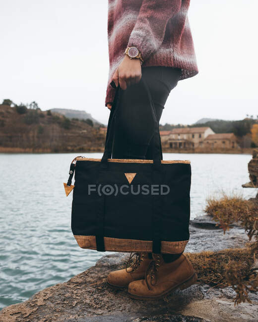 Side view of crop stylish female in casual outfit with trendy black handbag standing on riverbank in town in cloudy cold autumn day — Stock Photo