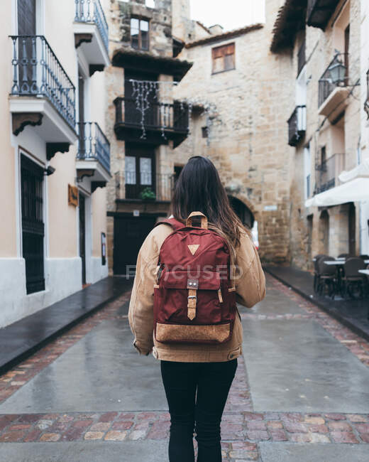 Back view of young female tourist in warm outerwear with trendy backpack walking on narrow street with old stone building in town — Stock Photo