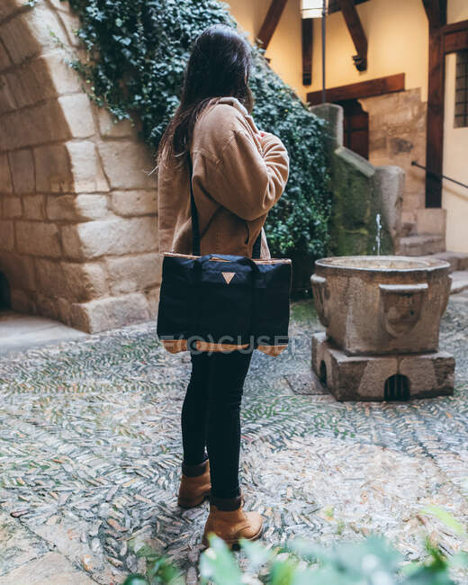 Side view of young female traveler in stylish outfit with bag on shoulder standing in yard of old stone building in autumn day in city — Stock Photo