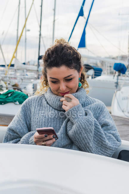 Jovem mulher em camisola casual sorrindo enquanto navega no celular no iate moderno — Fotografia de Stock