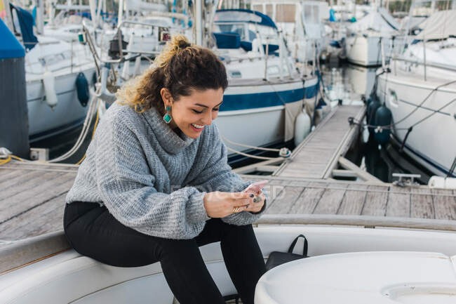 Молода жінка в повсякденному светрі посміхається, коли сидить на мобільному телефоні в сучасній яхті. — стокове фото