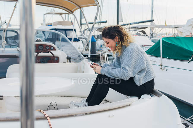 Vista lateral da jovem mulher em camisola casual sorrindo enquanto navega no celular no iate moderno — Fotografia de Stock