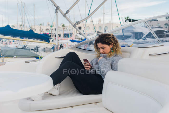 Vue latérale de la jeune femme en pull décontracté souriant tout en surfant sur téléphone mobile dans un yacht moderne — Photo de stock