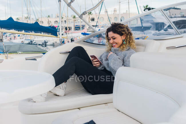 Вид збоку молодої жінки в повсякденному светрі посміхається під час серфінгу на мобільному телефоні в сучасній яхті — стокове фото