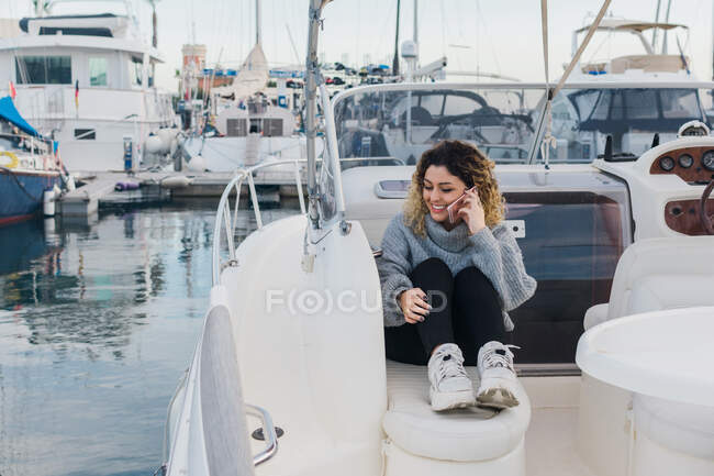 Mujer joven en suéter casual sonriendo mientras navega en el teléfono móvil en yate moderno - foto de stock