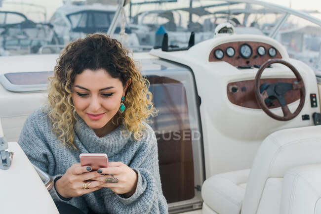 Dall'alto della giovane donna in maglione casual sorridente durante la navigazione sul telefono cellulare in yacht moderno — Foto stock