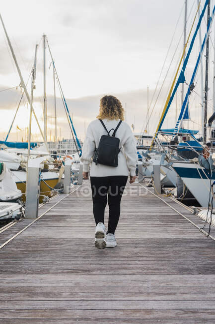 Вид на безликую молодую женщину в повседневной одежде с черным рюкзаком, идущую по деревянному пирсу между современными яхтами на закате — стоковое фото