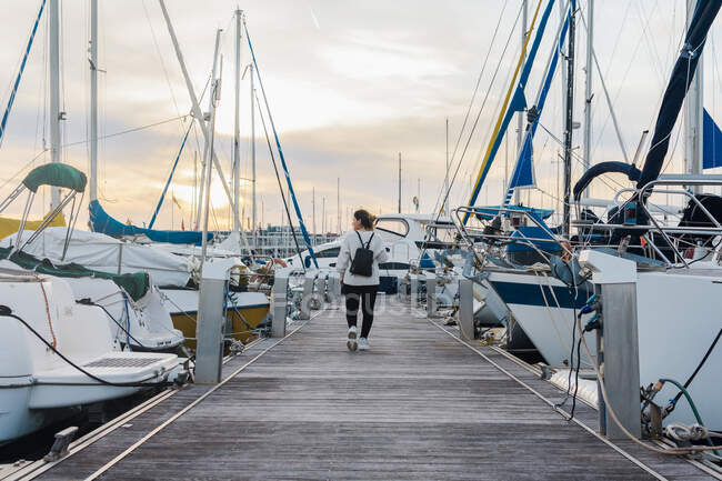 Back view of faceless young woman in casual wear with black backpack walking on wooden pier between modern yachts on sunset — Stock Photo