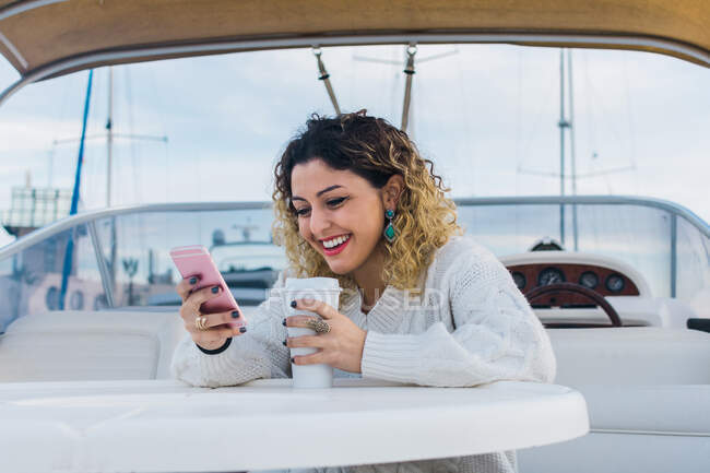 Молода жінка в повсякденному светрі посміхається під час серфінгу на мобільному телефоні в сучасній яхті — стокове фото