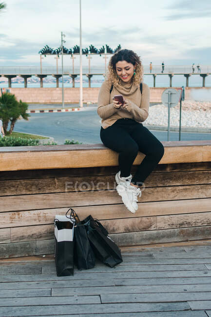 Zufriedene lockige junge Frau in lässiger Kleidung lächelt, während sie ihr Handy auf einer Bank mit Einkaufstaschen am modernen Kai benutzt — Stockfoto