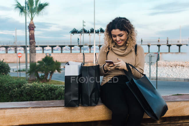 Mujer joven de pelo rizado satisfecho en ropa casual sonriendo mientras se utiliza el teléfono móvil en el banco con bolsas de compras en el muelle moderno - foto de stock