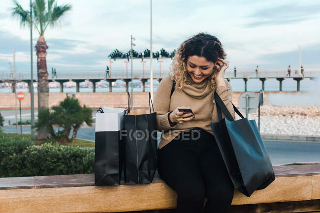 Satisfait jeune femme aux cheveux bouclés en tenue décontractée souriant tout en utilisant un téléphone mobile sur banc avec des sacs à provisions sur le quai moderne — Photo de stock