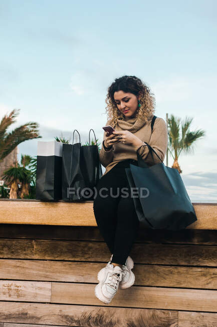 Soddisfatto giovane donna dai capelli ricci in abbigliamento casual sorridente durante l'utilizzo del telefono cellulare su panchina con borse della spesa su banchina moderna — Foto stock