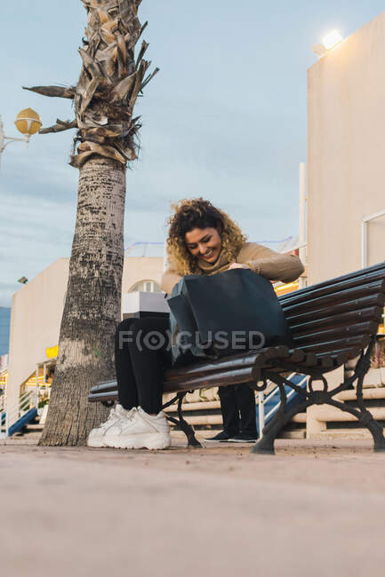 Side view from above of pleased curly haired lady smiling while checking shopping bar on wooden bench at city street — Stock Photo