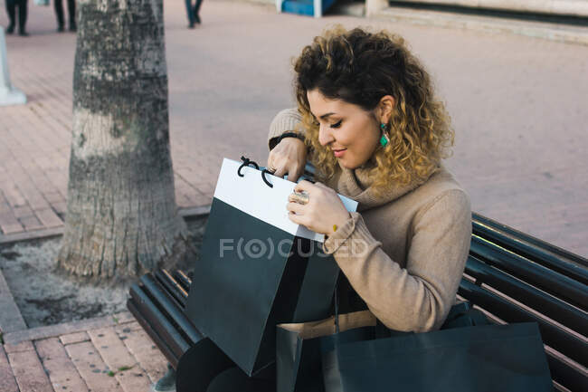 Vista lateral de cima da senhora de cabelos cacheados agradados sorrindo ao verificar o bar de compras no banco de madeira na rua da cidade — Fotografia de Stock