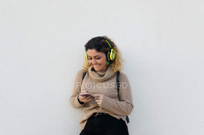Giovane donna felice in maglione casual beige sorridente durante l'utilizzo di cuffie e telefono cellulare con parete bianca sullo sfondo — Foto stock