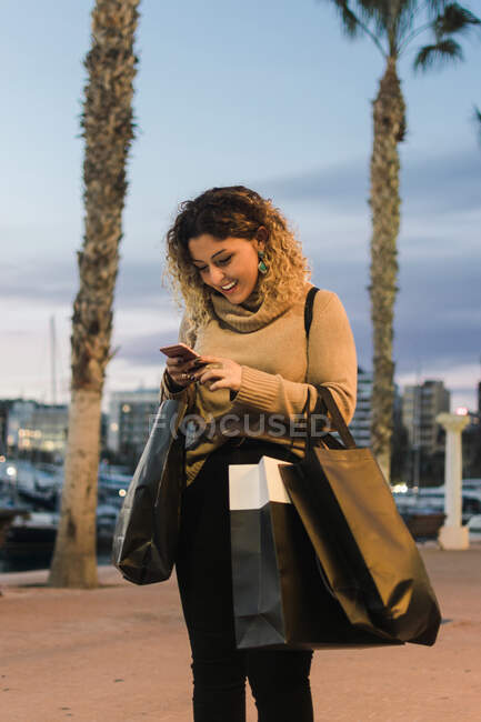 Vista lateral de la joven feliz con bolsas de compras riendo mientras envía mensajes de texto en el teléfono móvil con la ciudad moderna en el crepúsculo en el fondo - foto de stock