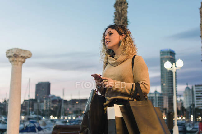 Vista laterale di felice giovane donna guardando lontano con borse della spesa ridere mentre messaggia sul telefono cellulare con la città moderna al crepuscolo sullo sfondo — Foto stock