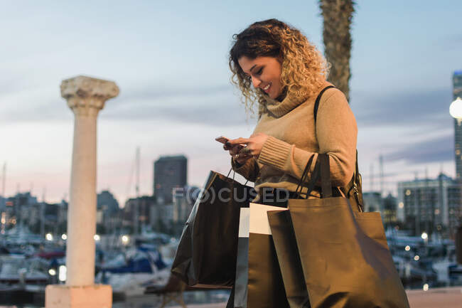 Seitenansicht einer glücklichen jungen Frau mit lachenden Einkaufstaschen beim SMS-Schreiben auf dem Handy mit moderner Stadt in der Dämmerung im Hintergrund — Stockfoto