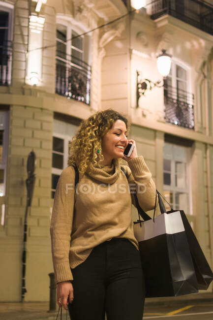 Jovencita alegre en ropa casual mirando hacia otro lado riendo mientras habla por teléfono móvil con bolsas de compras en la mano en la calle de la ciudad - foto de stock