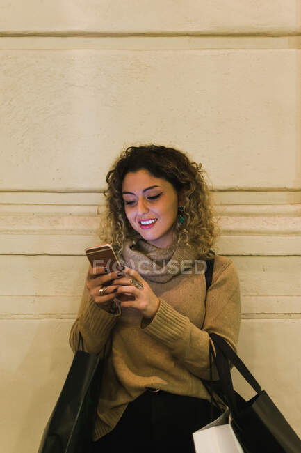 Задоволена кучерява молода жінка в повсякденному одязі посміхається, використовуючи мобільний телефон з сумками, що схиляються на стіні — стокове фото