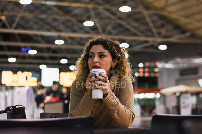 Современная леди в бежевом свитере, прислонившись локтями к столу с кофе, отправляется в аэропорт на размытом фоне — стоковое фото