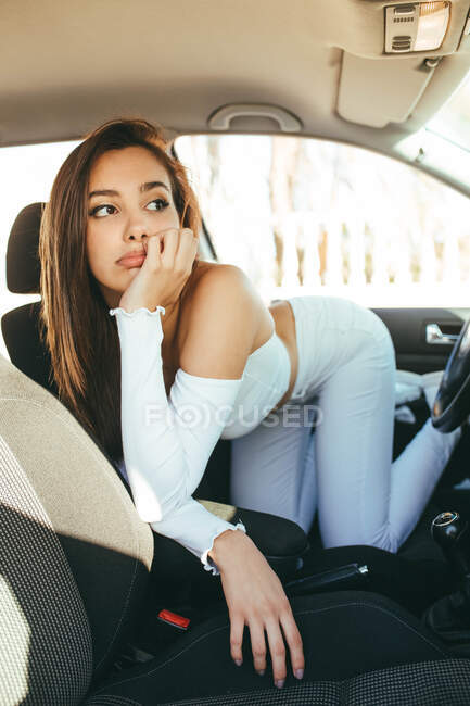 Скучная молодая чувствительная очаровательная женщина в повседневной одежде смотрит в сторону, стоя на коленях и опираясь на сиденье в машине — стоковое фото
