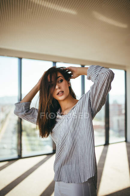 Вид ззаду на мрійливу чуттєву красиву жінку в смугастій сорочці, що показує неозброєне плече, стоячи в кімнаті з панорамними вікнами — стокове фото